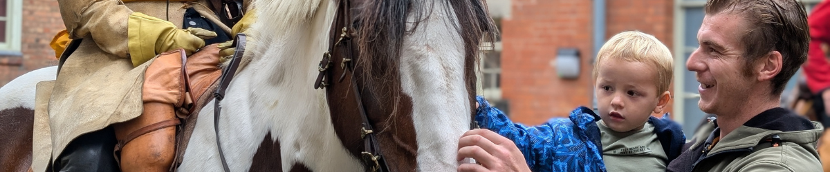 Image of a child being carried by male figure, stroking a horse